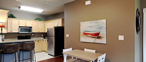 A modern kitchen with wooden cabinets, a stainless steel refrigerator, a microwave, and a ceiling fan. There is a dining area with a wooden table and white chairs.