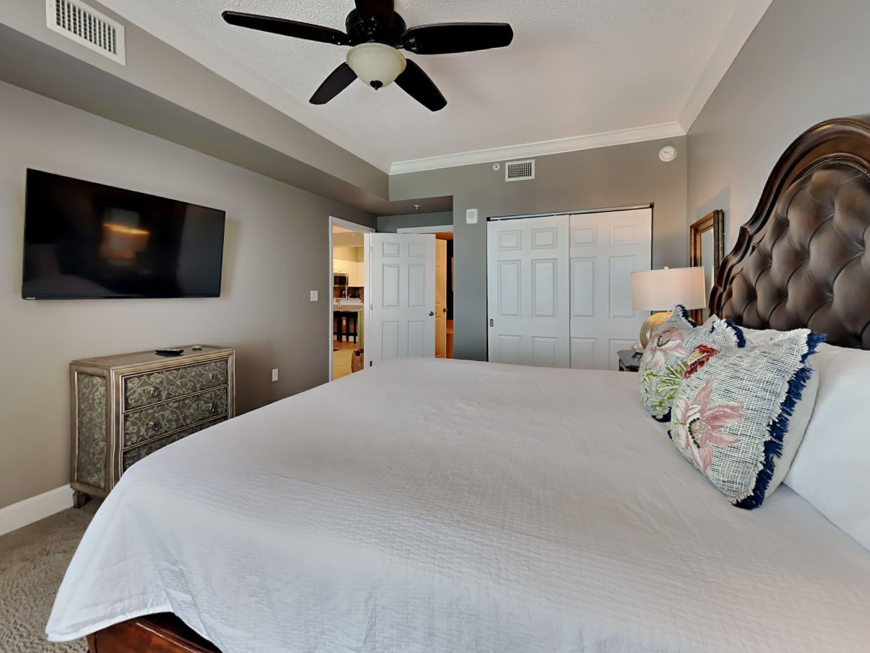 A bedroom with a large bed, a ceiling fan, a TV, and a dresser.