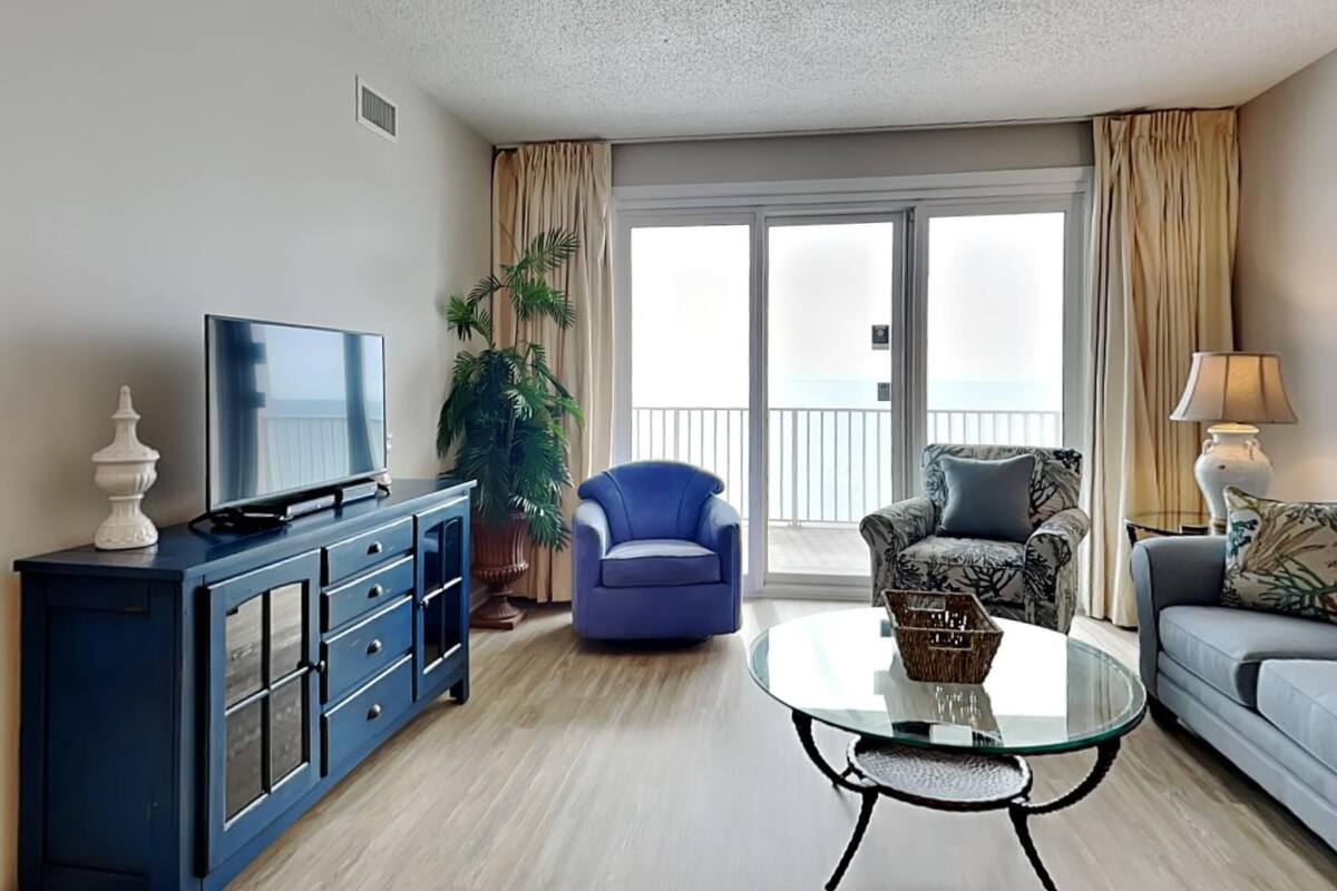 A modern living room with a flat-screen TV, a blue armchair, a patterned sofa, a glass coffee table, and a large window with a view of the sea.