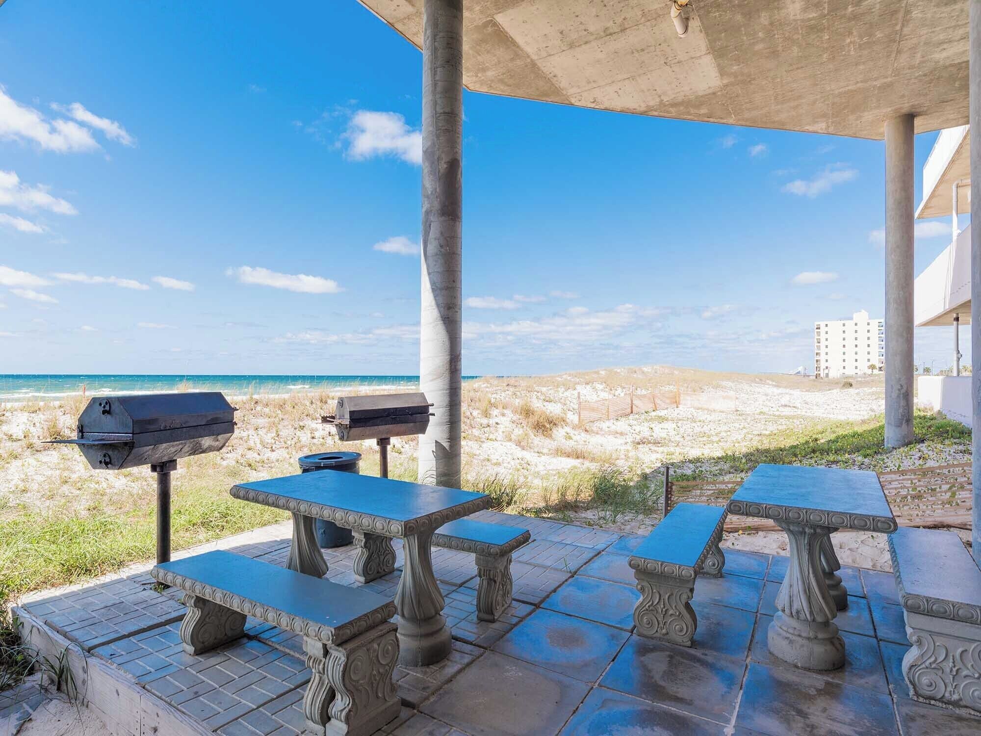 An outdoor patio with stone tables and a barbecue grill, overlooking a beach and ocean.