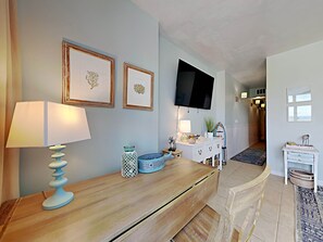 Una sala de estar con una mesa de centro de madera, una lámpara, obras de arte enmarcadas, un televisor y un armario blanco.