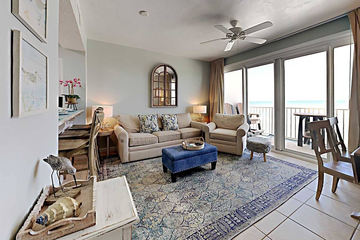 A spacious living room with a large window overlooking the ocean, a ceiling fan, and a balcony with outdoor seating.