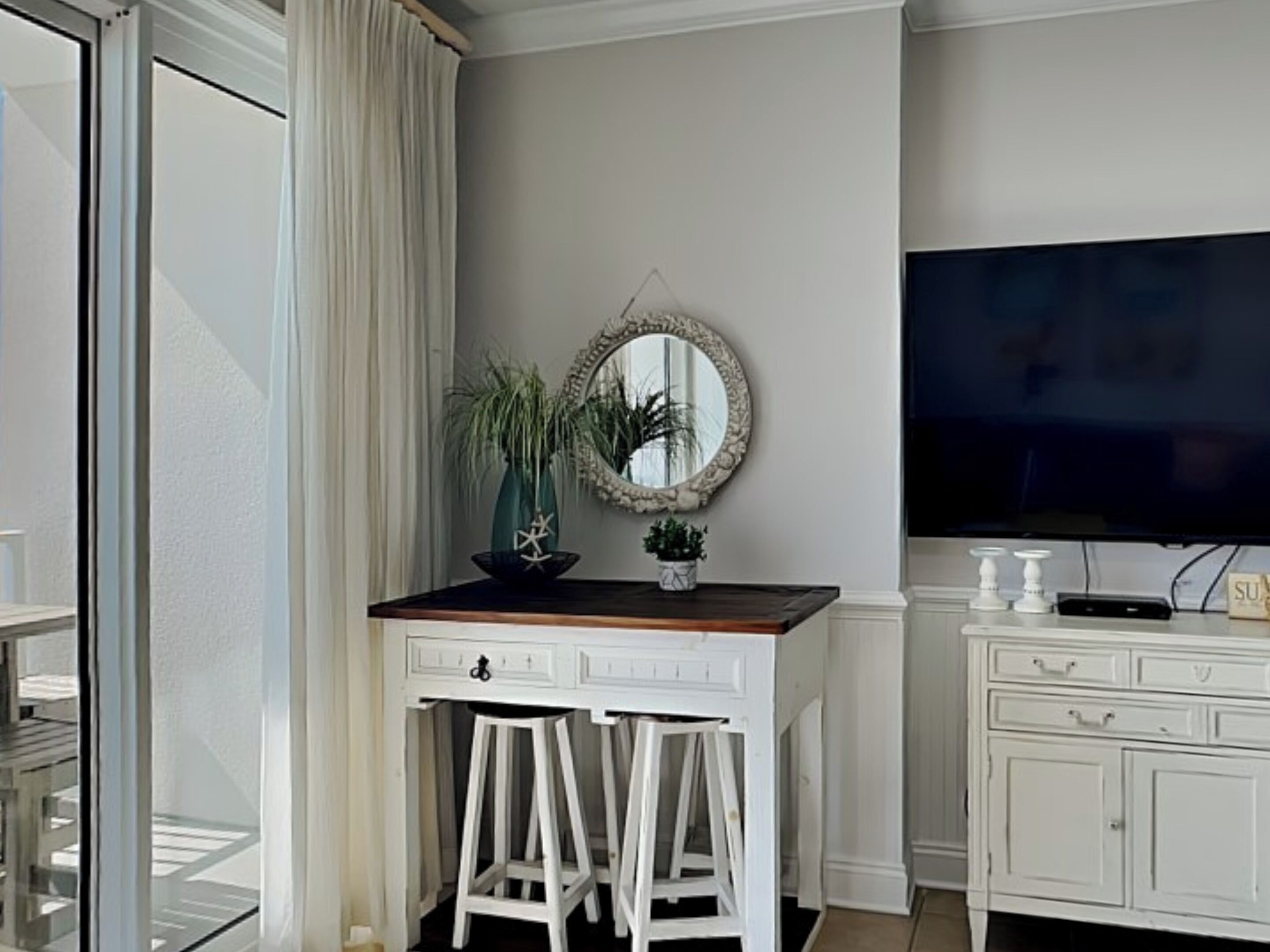 A room with a white desk, two white stools, a mirror, a television, and a cabinet.