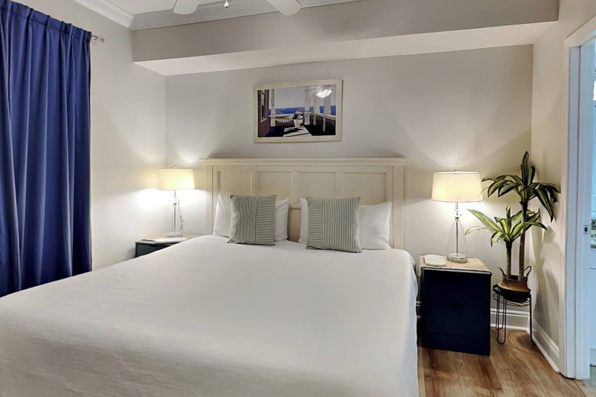 A hotel room with a large bed, two bedside tables, a ceiling fan, a framed picture on the wall, and a potted plant.