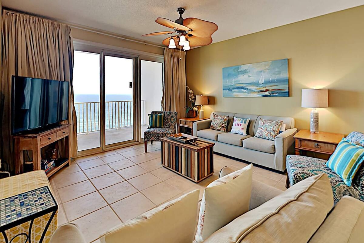 A living room with a television, sofa, and a table. There is a balcony with a view of the sea.