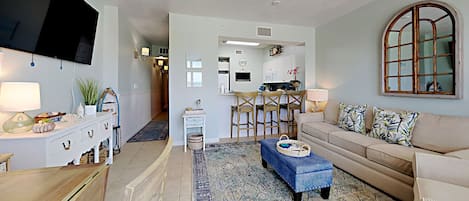 A living room with a TV, sofa, and coffee table. There is a kitchen in the background with a dining area.