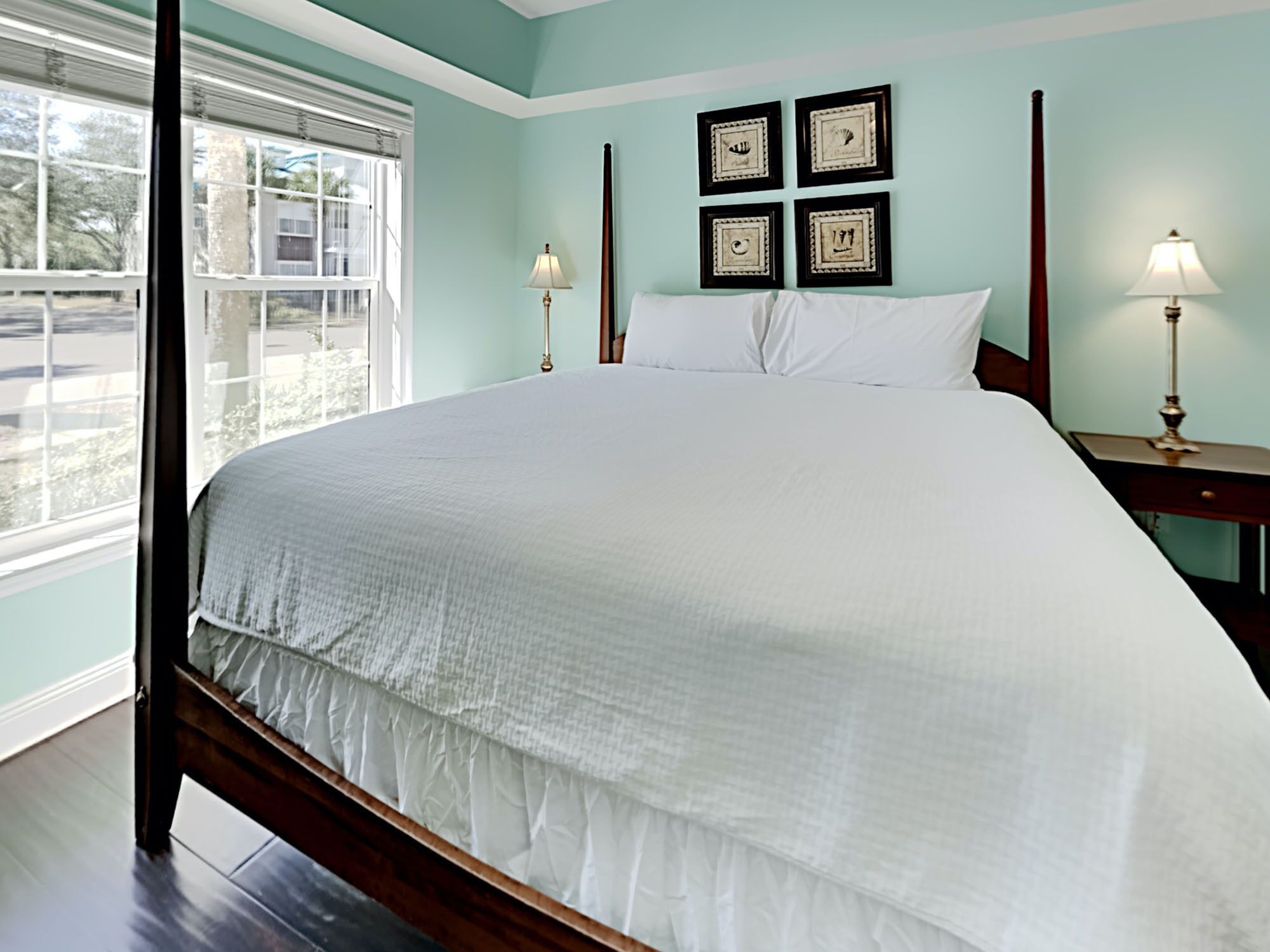 A neatly made bed with a white comforter and pillows, a wooden headboard, and a nightstand with a lamp. There are four framed pictures on the wall above the bed.