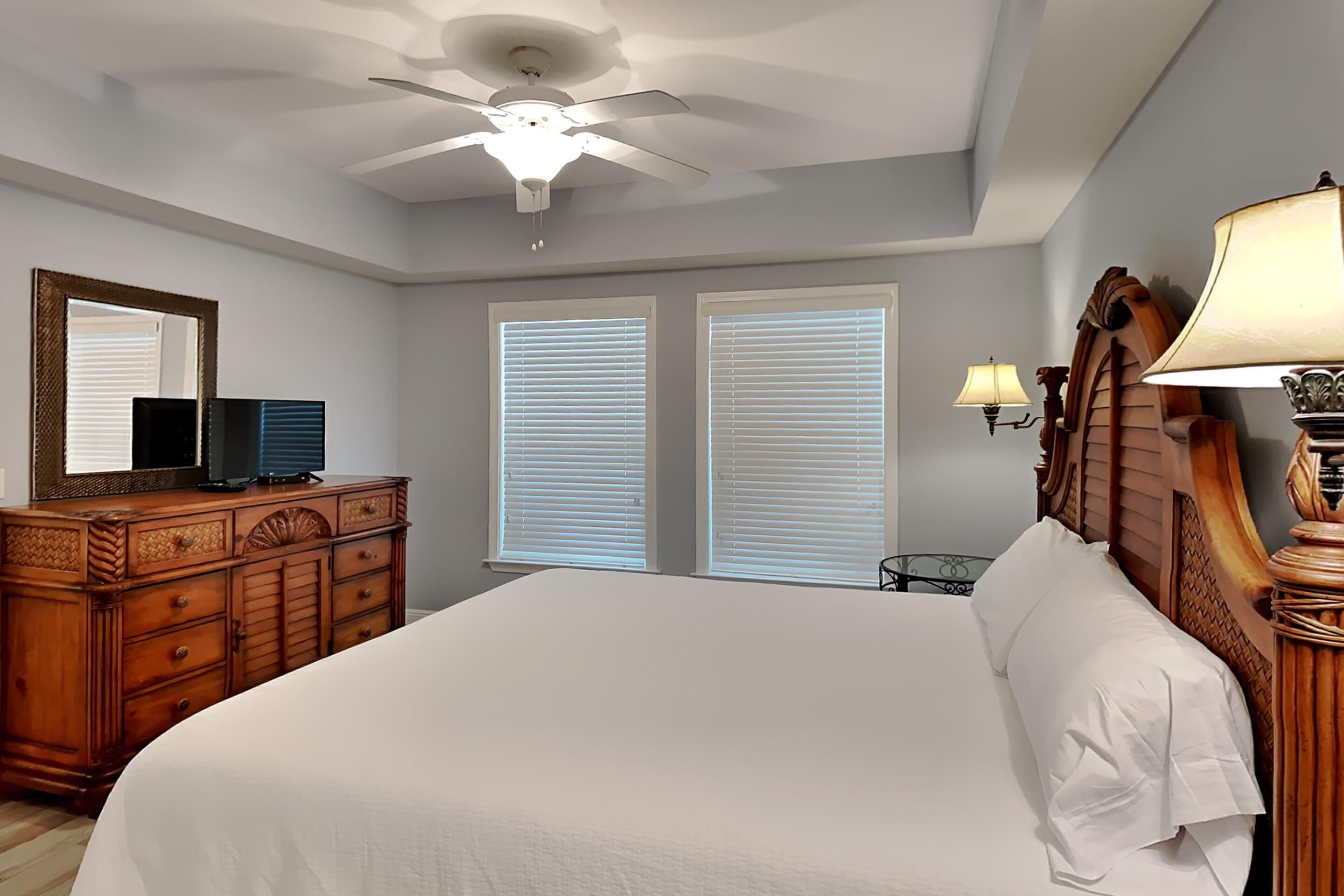 A bedroom with a large bed, wooden dresser, ceiling fan, two windows with blinds, and a nightstand with a lamp.