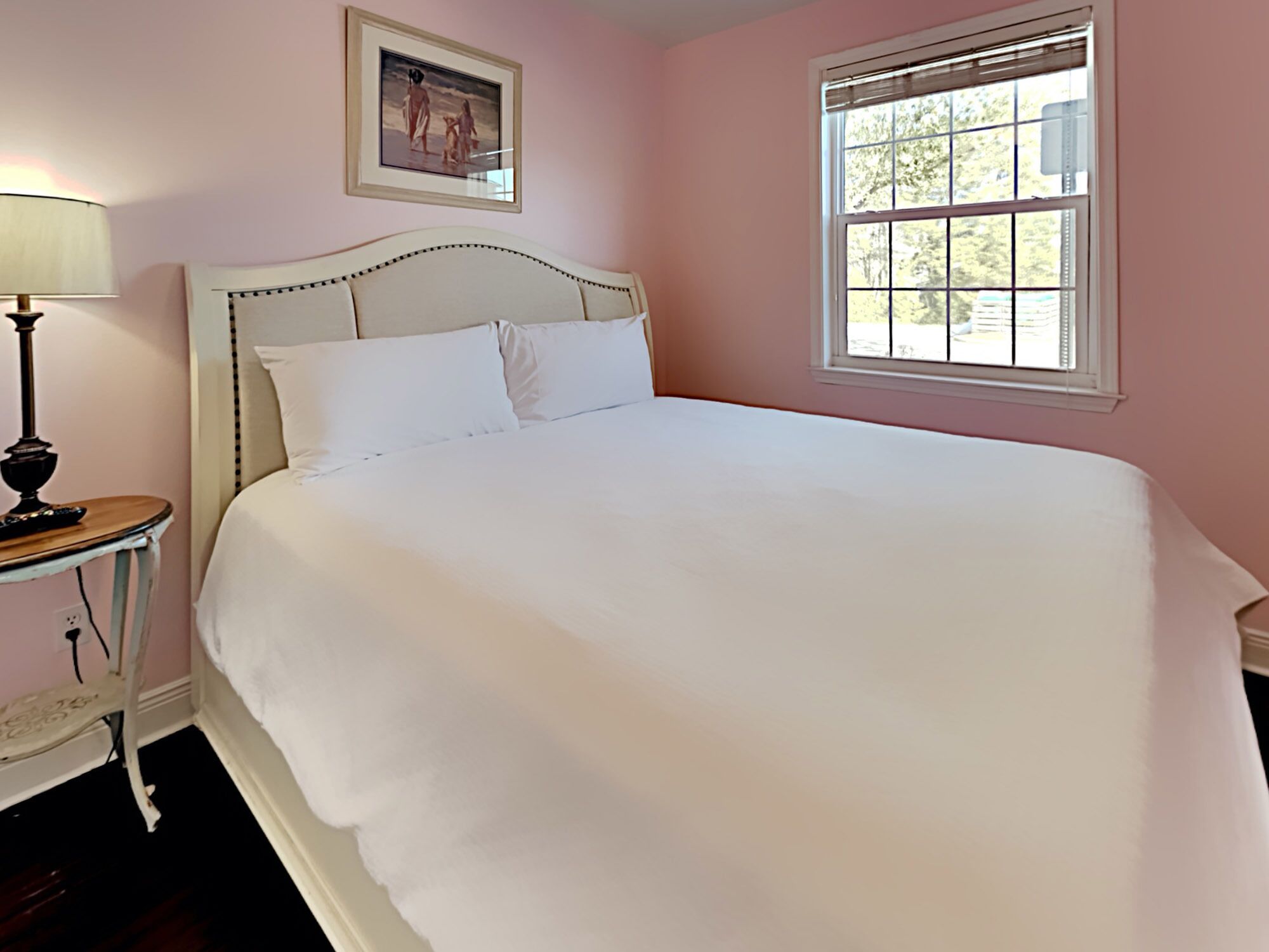 A bedroom with a large bed, a bedside table with a lamp, a window with blinds, and a framed picture on the wall.
