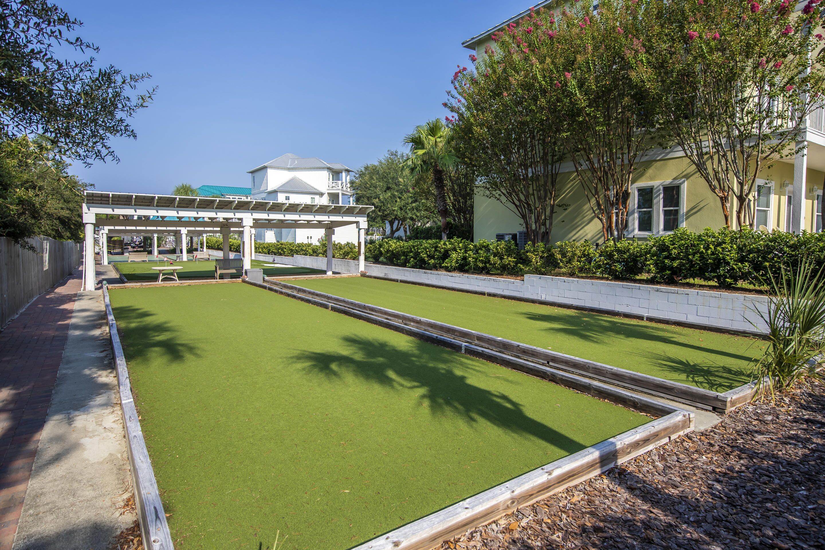 A green artificial lawn with a wooden pergola structure, a white building with a balcony in the background, and a tree with pink flowers to the right.