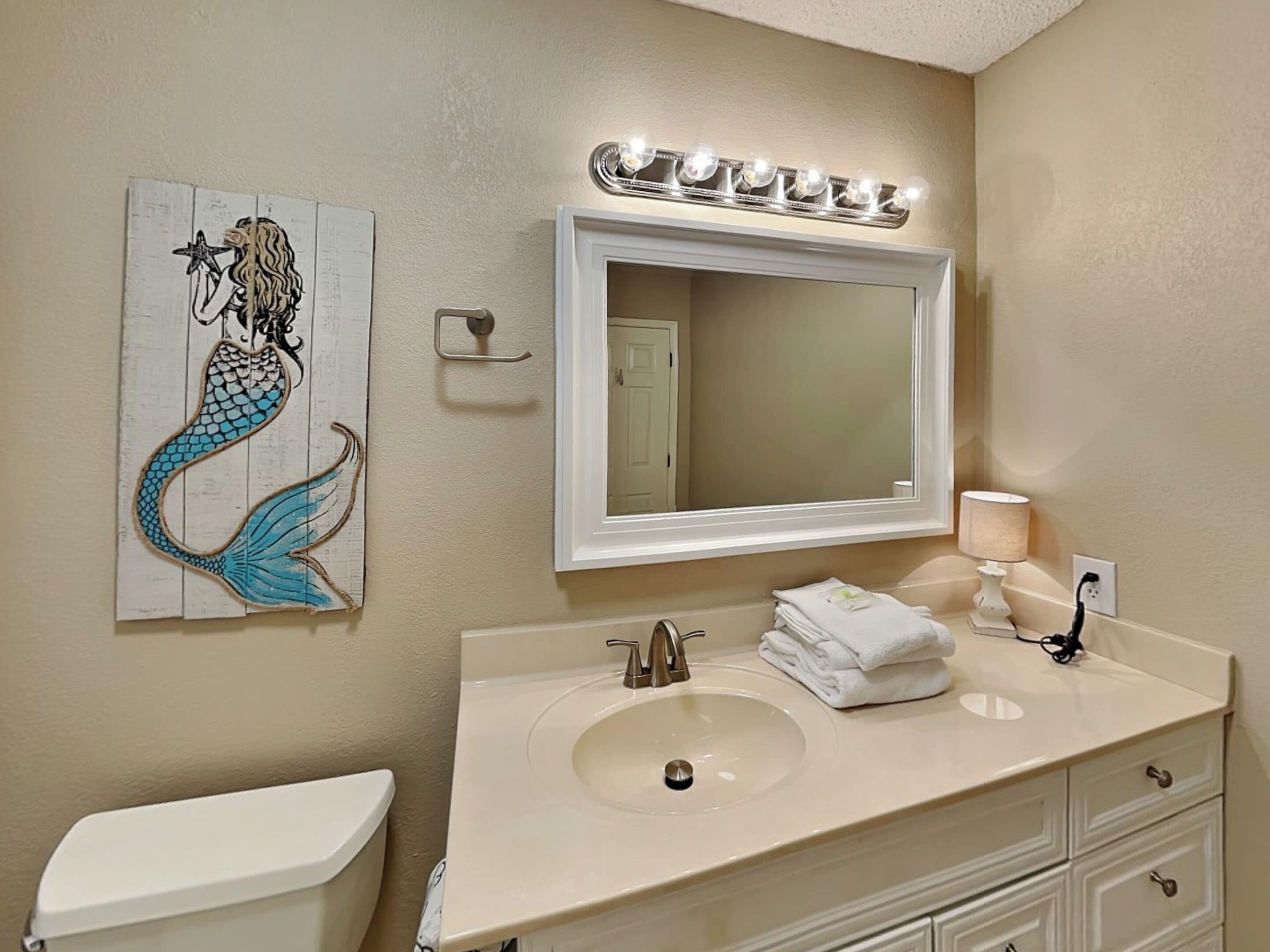Een badkamer met een wit wastafelmeubel, een grote spiegel en een kunstwerk van een zeemeermin aan de muur.
