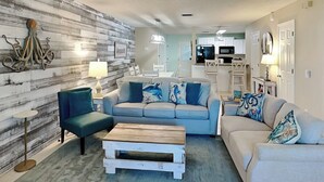 Un salon avec un canapé bleu, un fauteuil turquoise et une table basse en bois. On peut voir un ventilateur de plafond au-dessus.