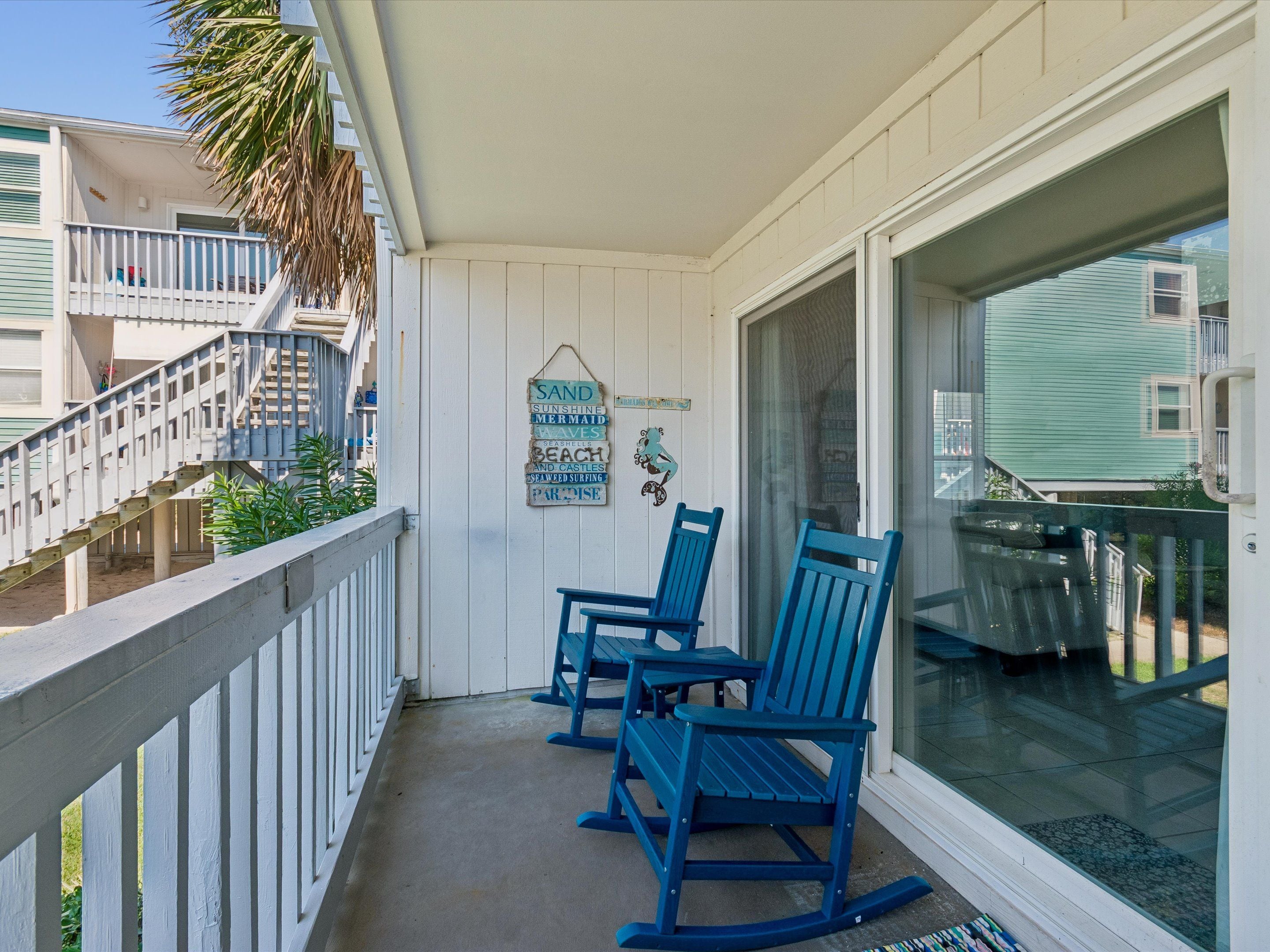 A balcony with two rocking chairs, a palm tree, and a view of neighboring houses.