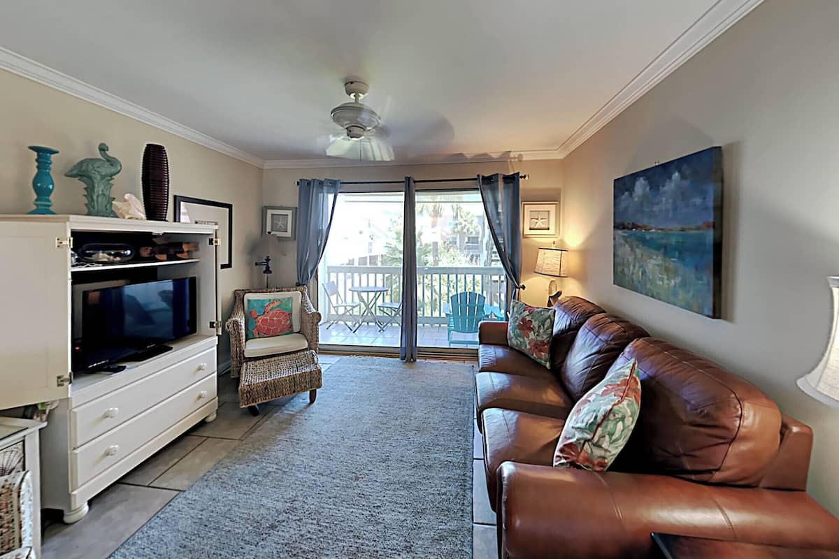 A living room with a television, a brown leather sofa, a wicker chair, and a balcony with outdoor furniture.