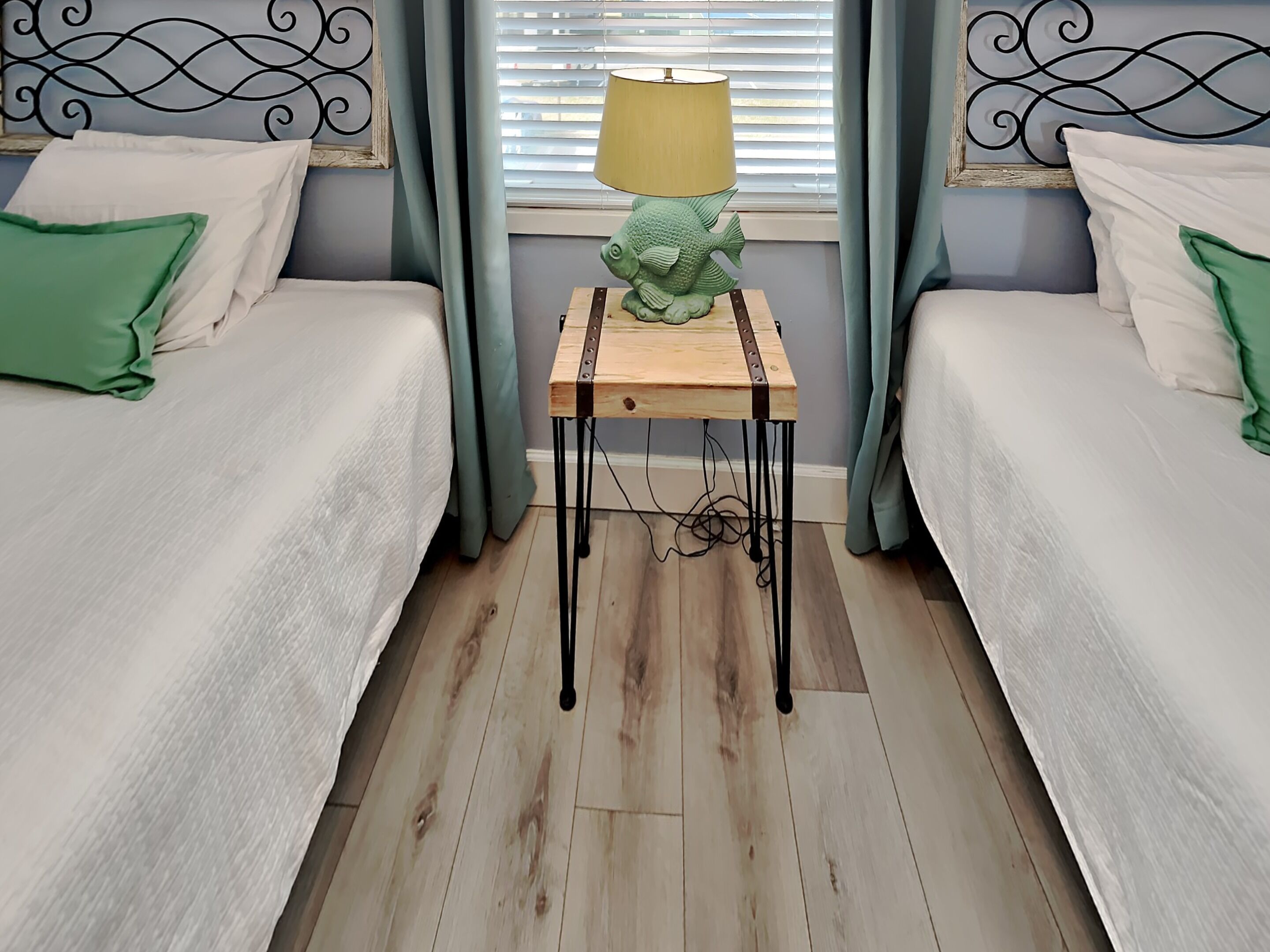 A bedroom with two beds, a wooden nightstand with a lamp, and a window with blinds.