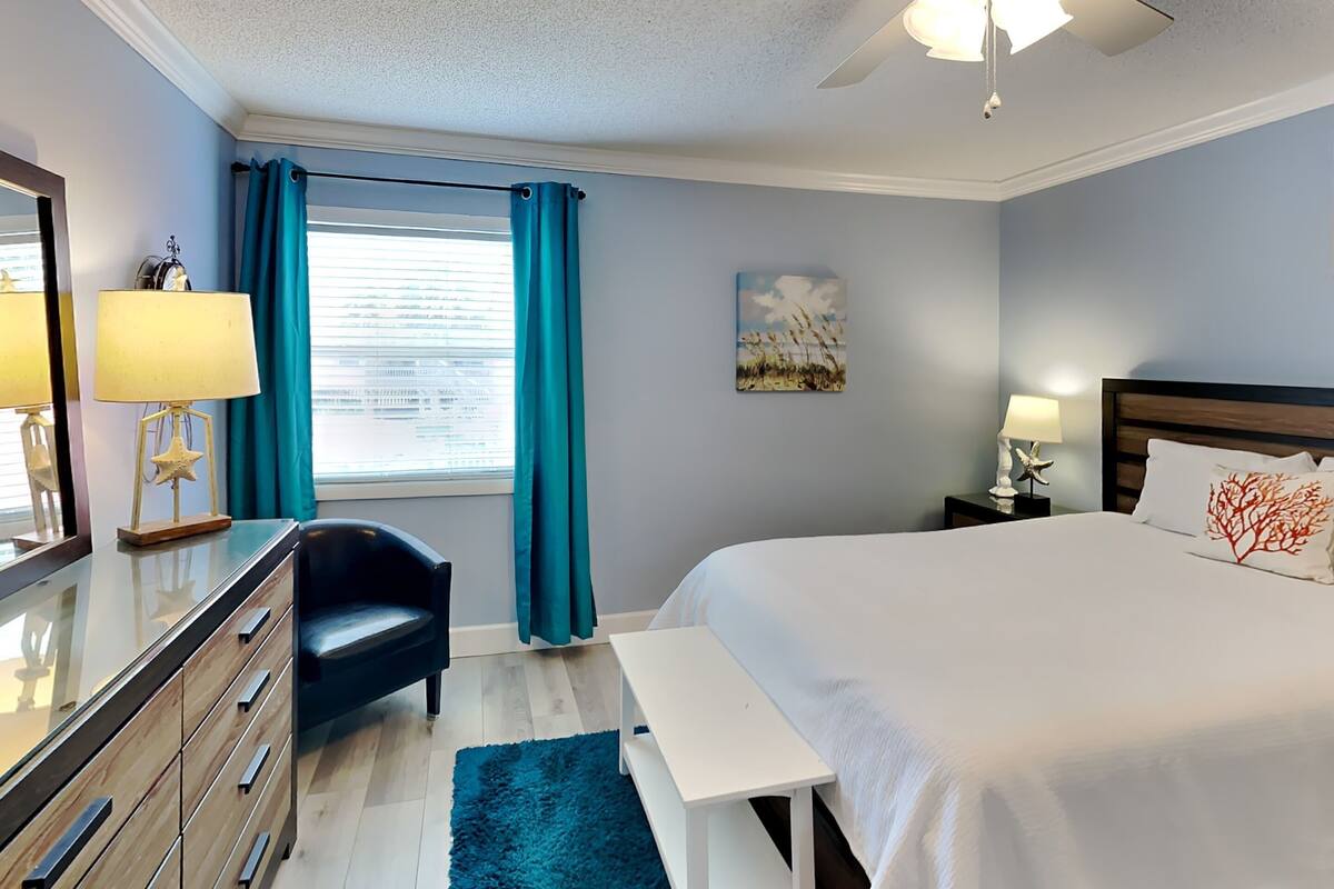 A bedroom with a bed, a desk, a chair, a nightstand, a lamp, a ceiling fan, and a window with curtains.