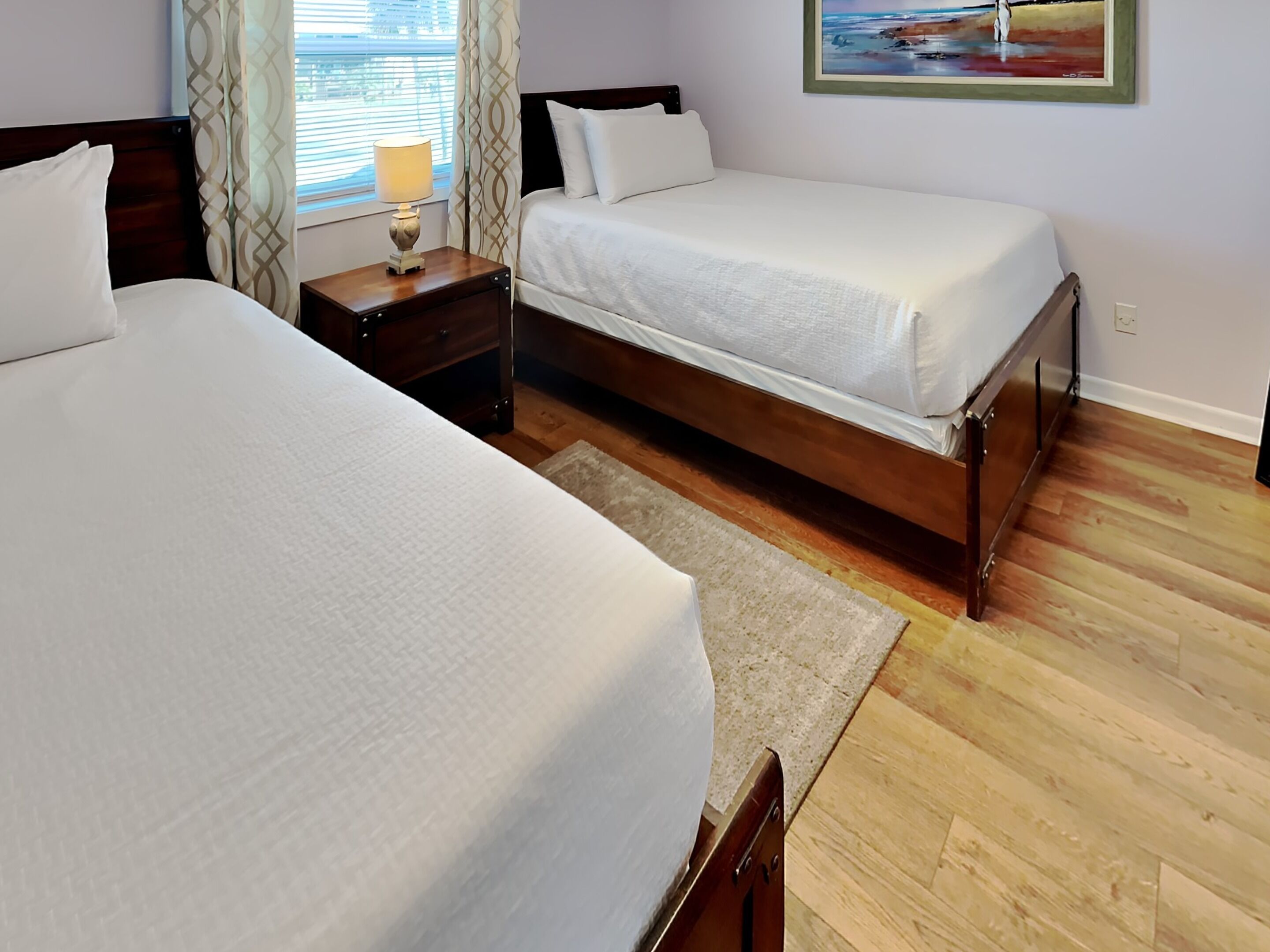 A hotel room with two beds, a nightstand with a lamp, a painting on the wall, and wooden flooring.