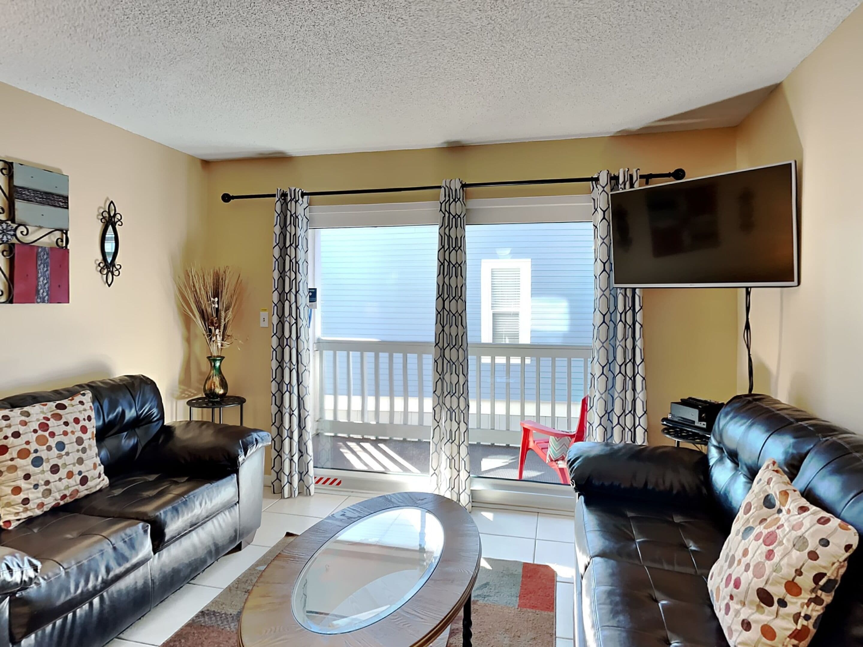 A living room with two leather sofas, a glass coffee table, a flat-screen TV, and a balcony with a view.