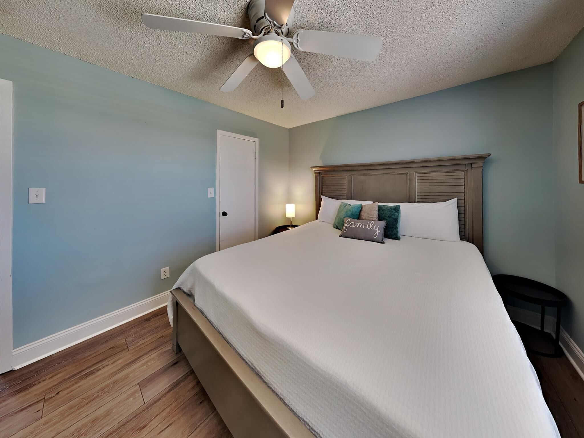 A bedroom with a large bed, a ceiling fan, a door, and a nightstand with a lamp.