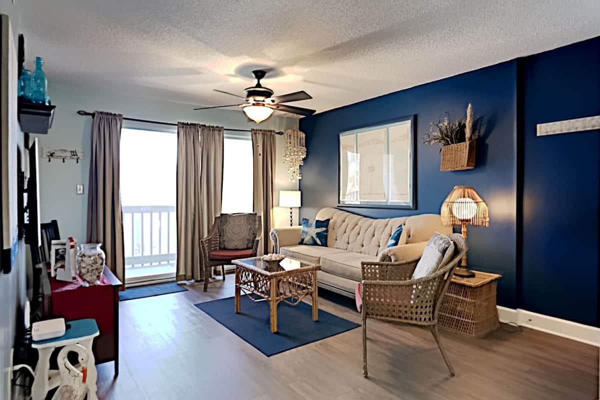 Un salon avec un mur d’accent bleu, un ventilateur de plafond, un canapé avec des coussins, une table basse et un fauteuil en osier.