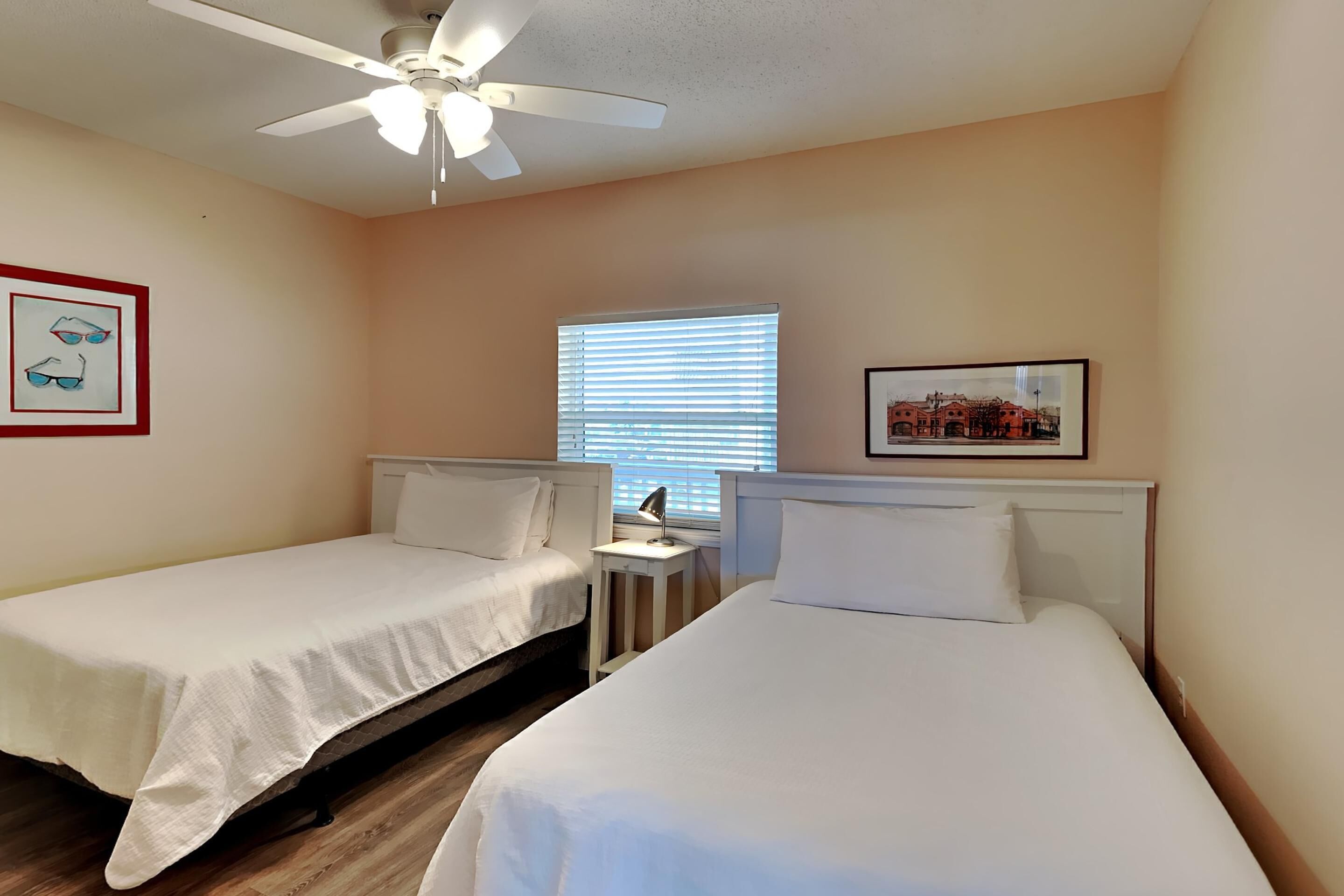 A hotel room with two single beds, a ceiling fan, a window with blinds, and framed pictures on the walls.