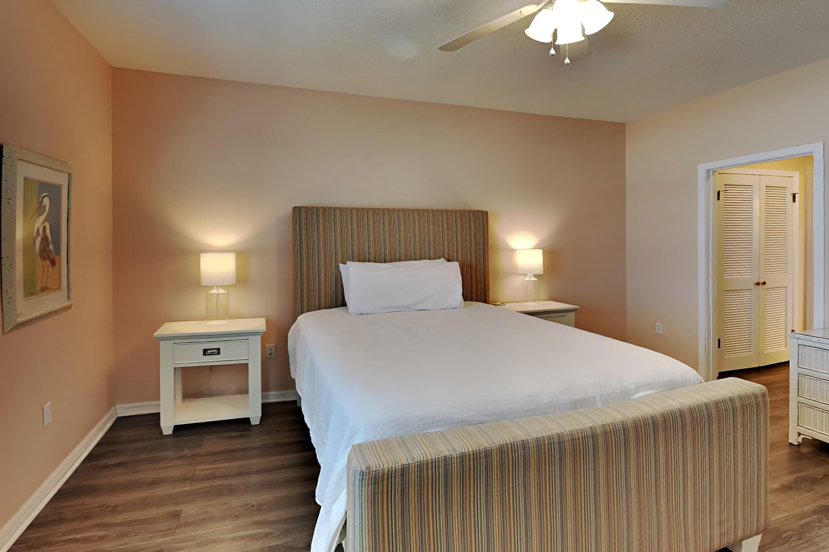 Une chambre à coucher bien aménagée, avec un grand lit, des tables de chevet avec des lampes, un ventilateur de plafond et une porte dotée de volets à persiennes.
