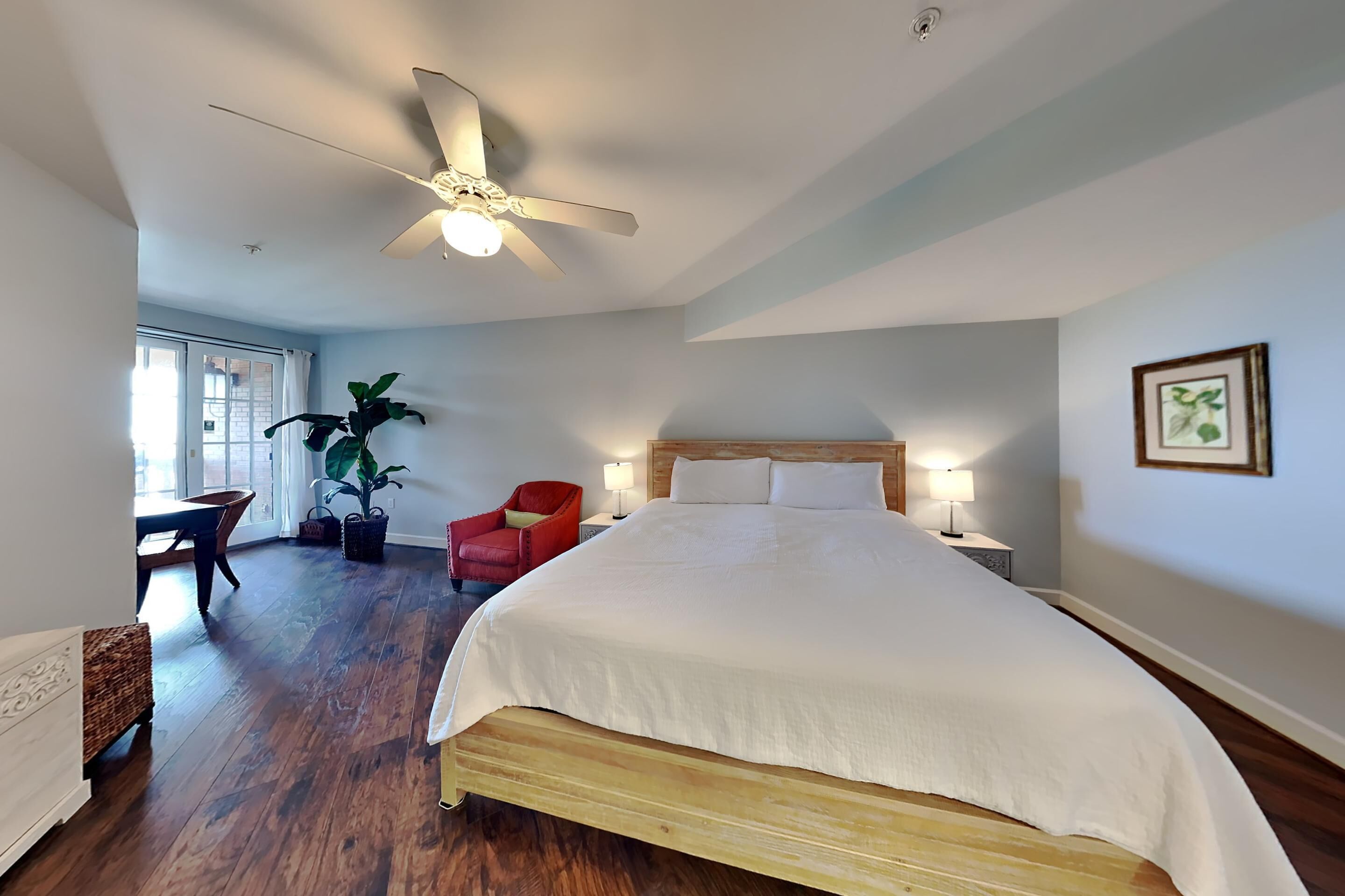 Une chambre avec un grand lit, un ventilateur de plafond, une chaise rouge, une table et une plante en pot.
