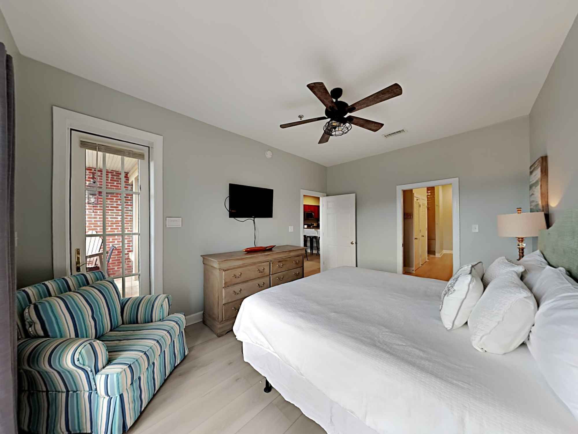 Une chambre à coucher comprenant un lit, un téléviseur, un ventilateur de plafond, une commode et un fauteuil à rayures.