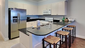 A modern kitchen with white cabinets, a stainless steel refrigerator, a microwave, and a stove. There is a black countertop with a sink and bar stools.