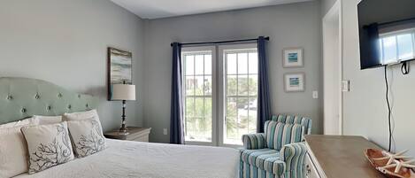A bedroom with a bed, a chair, a dresser, a ceiling fan, a window with curtains, and artwork on the walls.