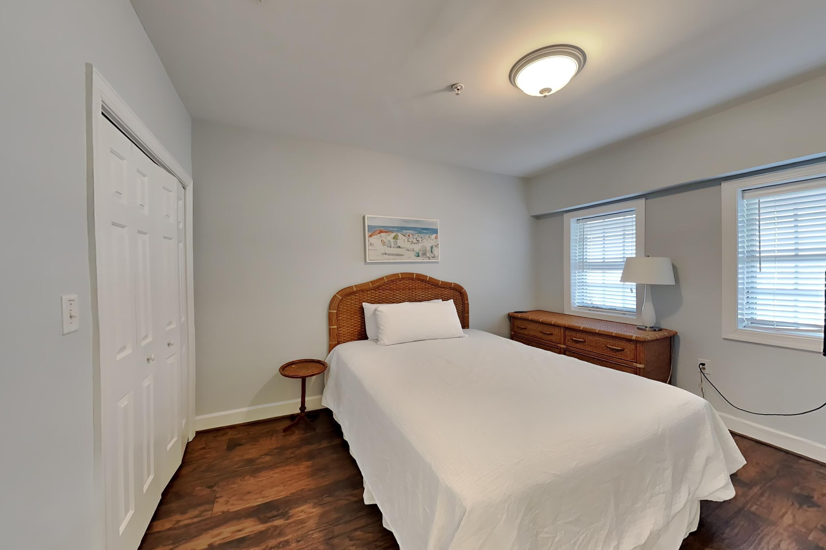 A bedroom with a large bed, a wooden dresser, a nightstand, a wall-mounted lamp, and a painting on the wall.