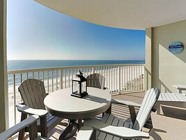 A balcony with a round table and four Adirondack chairs, overlooking a beach with the ocean in the background.
