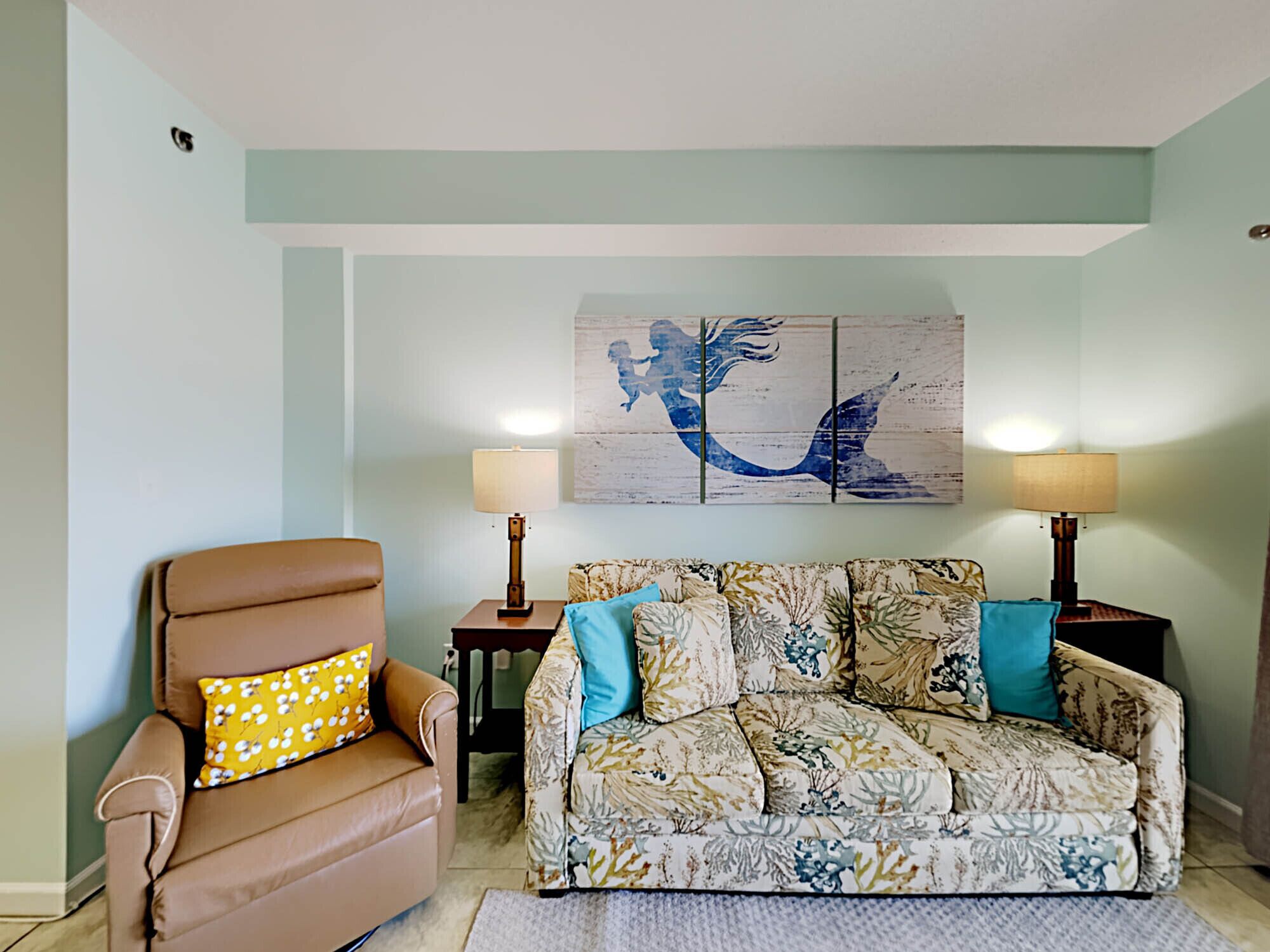 A living room with a brown leather armchair, a floral-patterned sofa, two side tables with lamps, and a wall-mounted artwork of a mermaid.