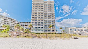 Ein Strandhotel mit mehreren Balkonen, ein Sandstrand im Vordergrund und Palmen.