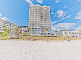 A beachfront hotel with multiple balconies, a sandy beach in the foreground, and palm trees.