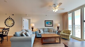A living room with a sofa set, a coffee table, a chair, and a view of the city through large windows. - Waterscape by Southern Vacation Rentals (Fort Walton Beach)