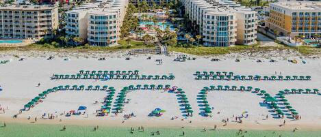 Un hotel frente a la playa con varias edificaciones, una piscina grande y una playa con sombrillas dispuestas en forma de corazón.