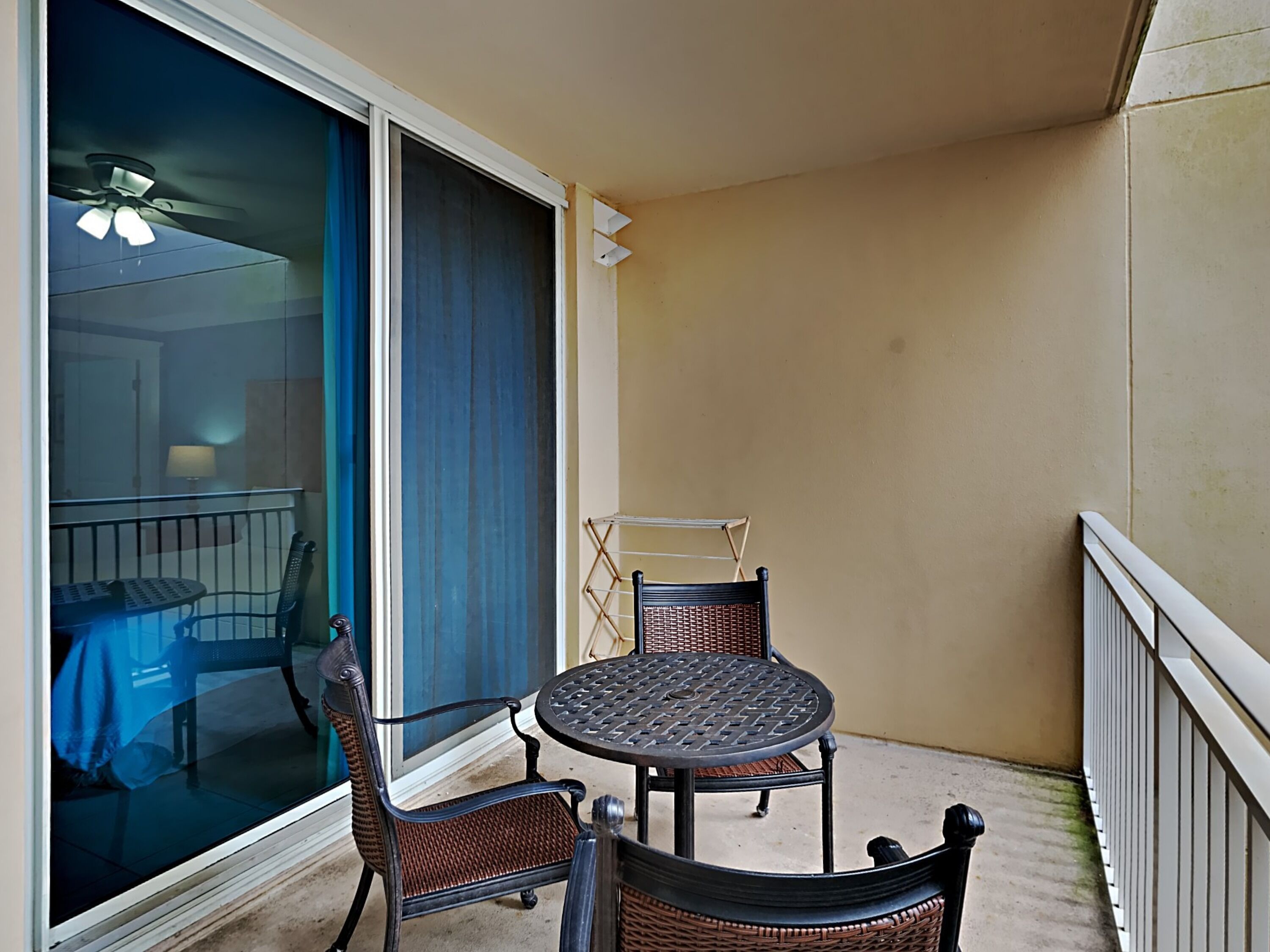A balcony with a round table and two wicker chairs, a ceiling fan, and a view of a room with a table and chairs.