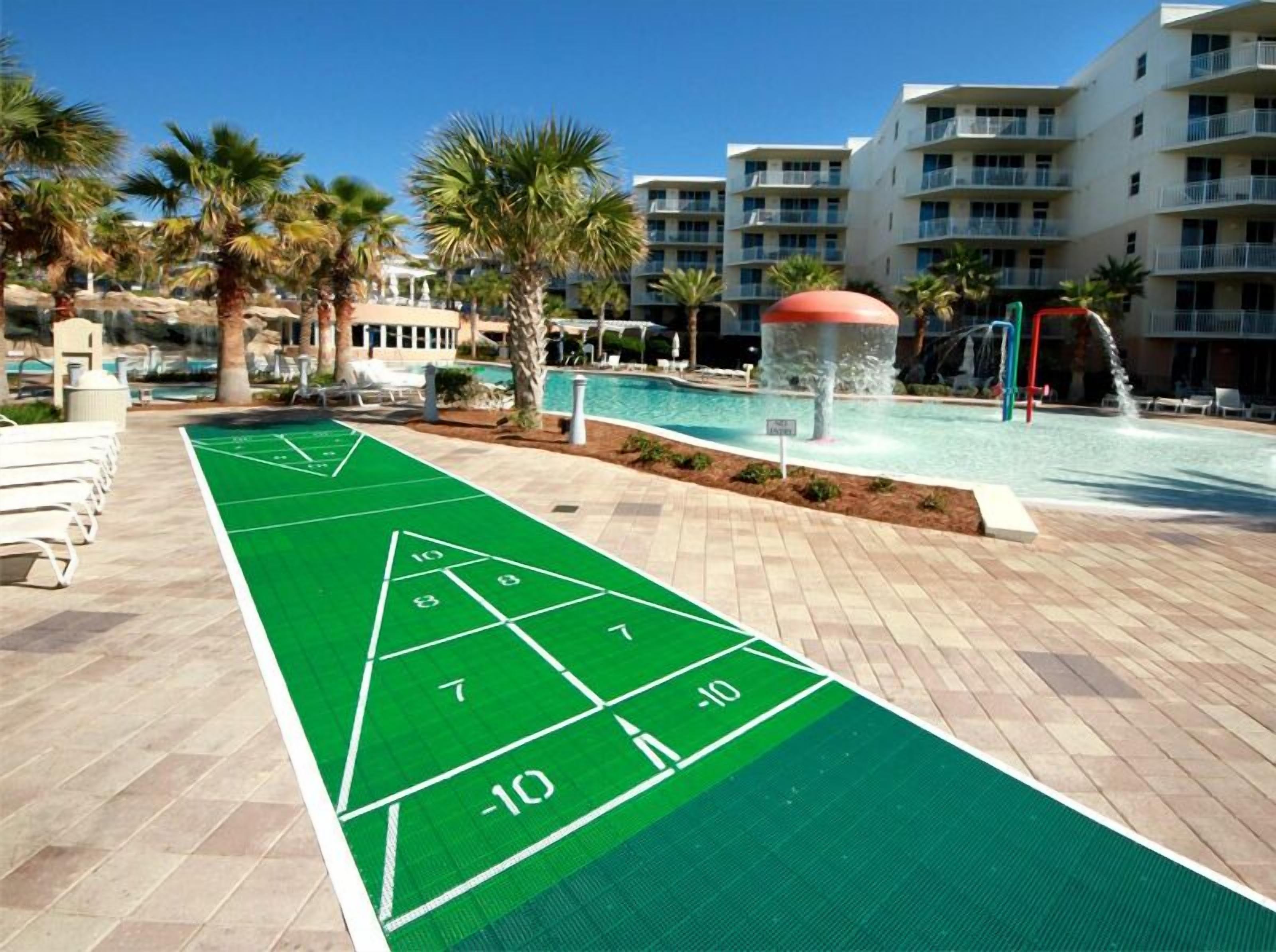 A pool area with a shuffleboard court, lounge chairs, palm trees, and a water fountain.