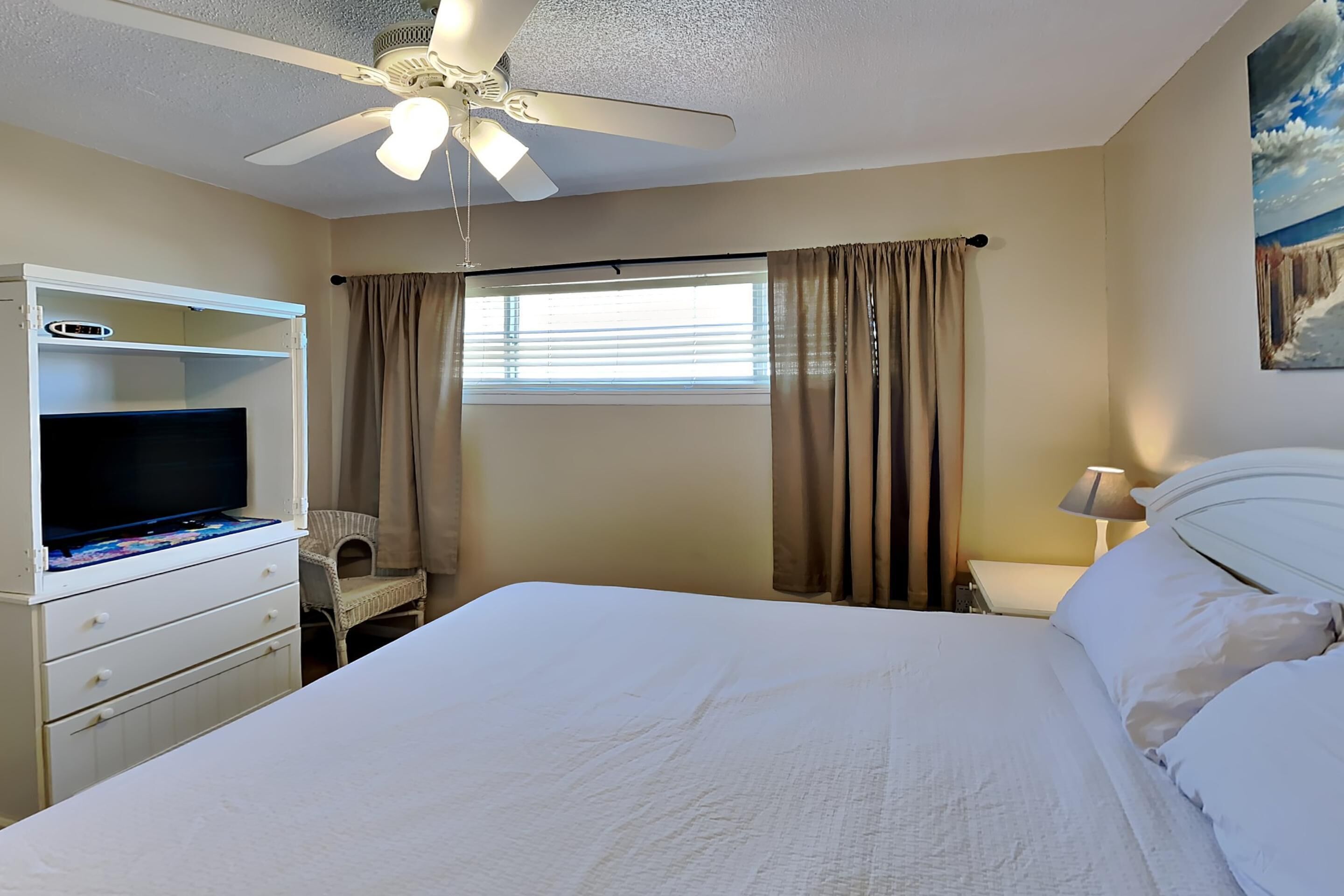 Une chambre à coucher avec un lit, un téléviseur posé sur une commode, un ventilateur de plafond, une fenêtre avec des rideaux et un tableau accroché au mur.