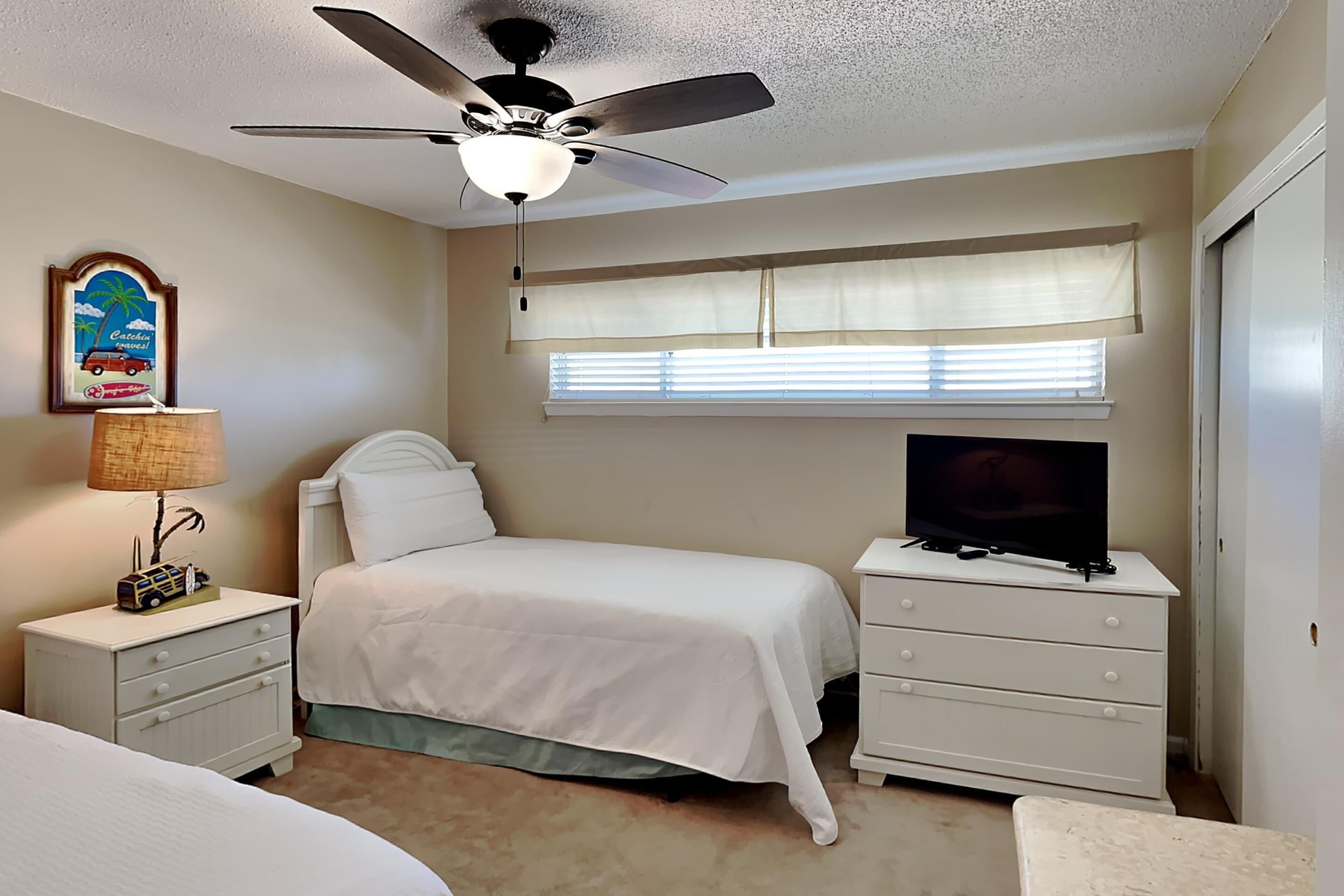 Une chambre à coucher comprenant un lit, une table de chevet avec une lampe, une commode, un ventilateur de plafond, un téléviseur et une fenêtre avec des stores.