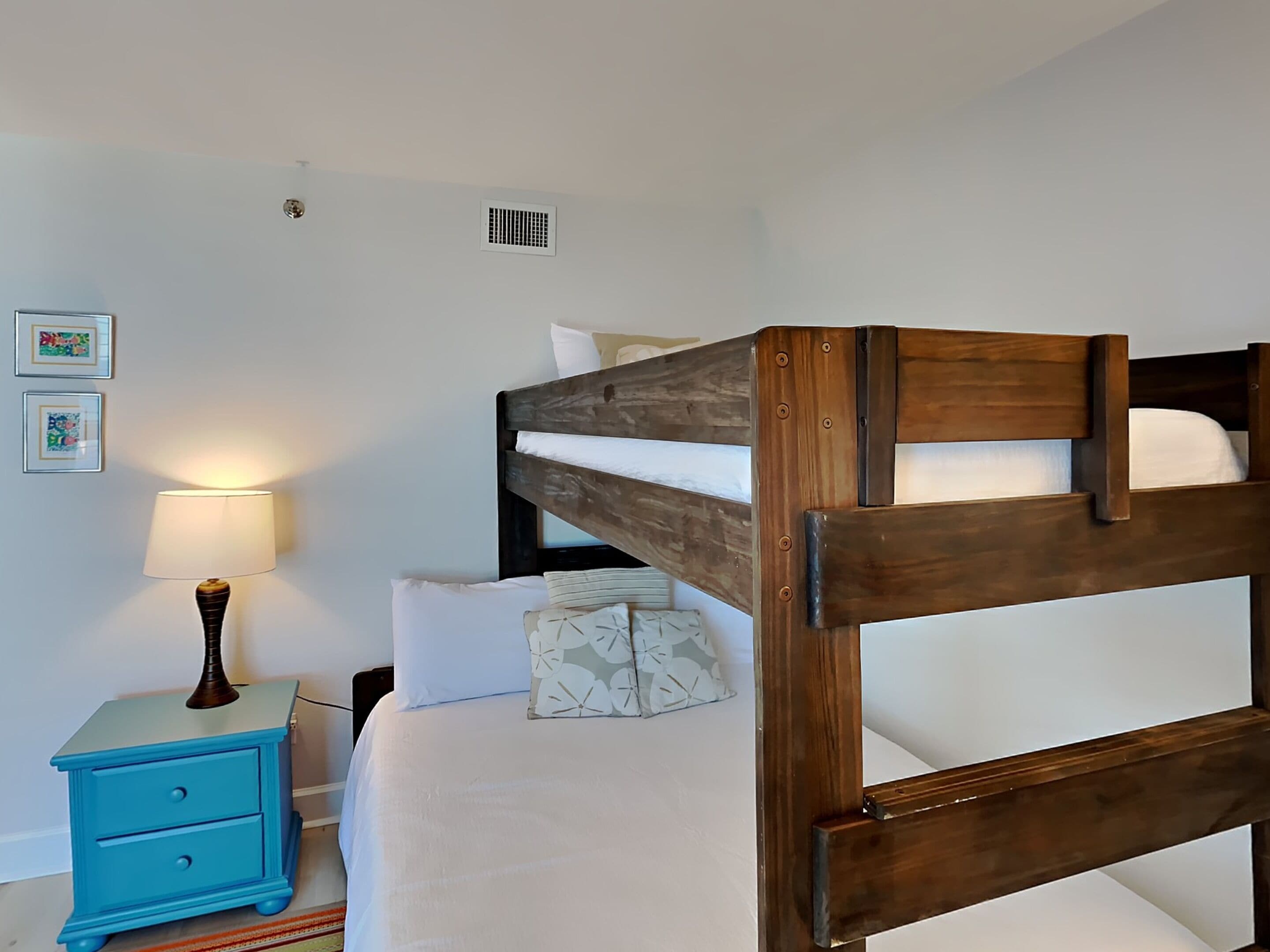 Una cama litera con estructura de madera, una mesita de noche con lámpara y dos cuadros enmarcados en la pared.