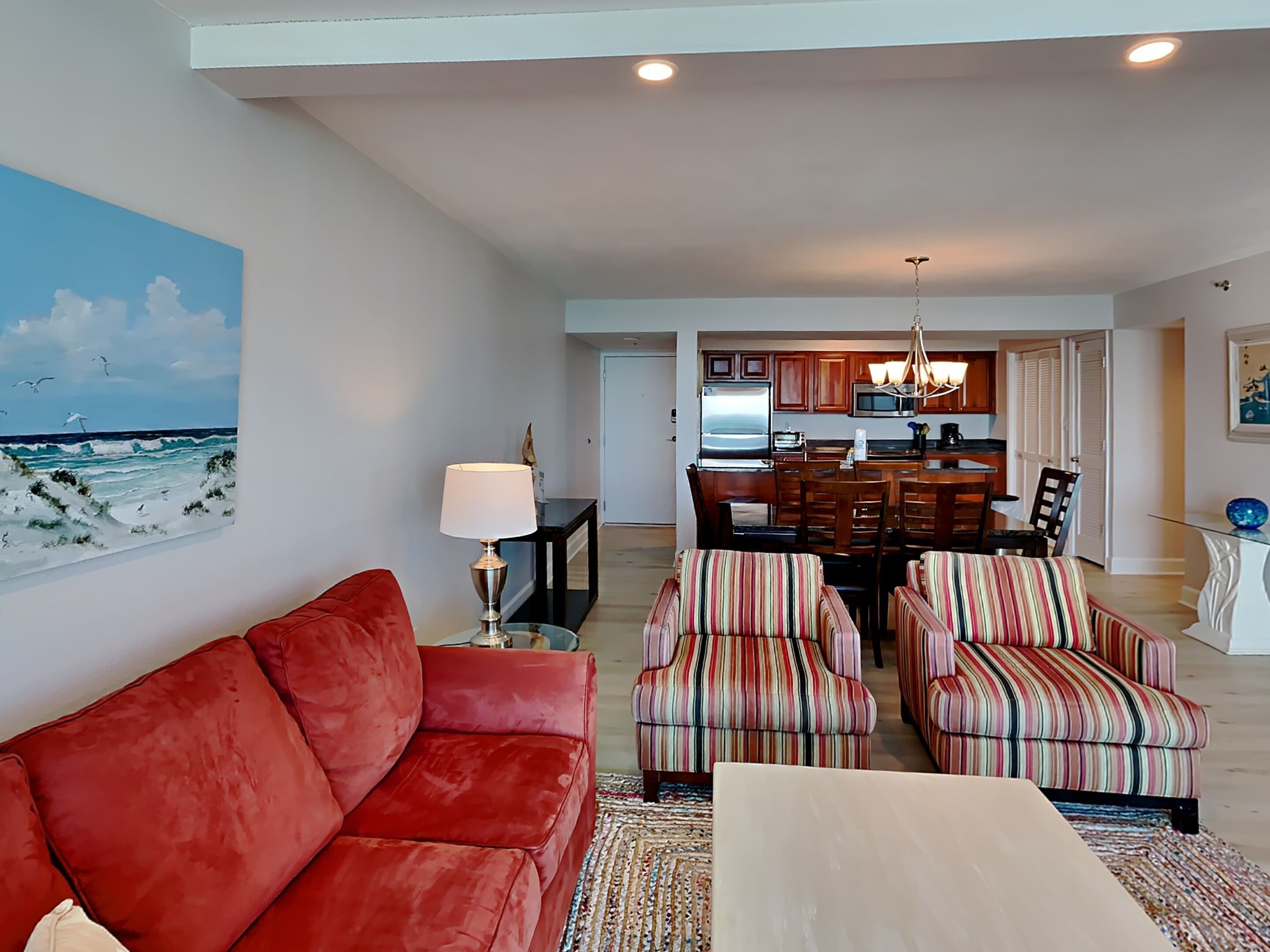 Ein Wohnzimmer mit einem roten Sofa, gestreiften Sesseln, einem Couchtisch und einem Gemälde, das eine Strandansicht zeigt.