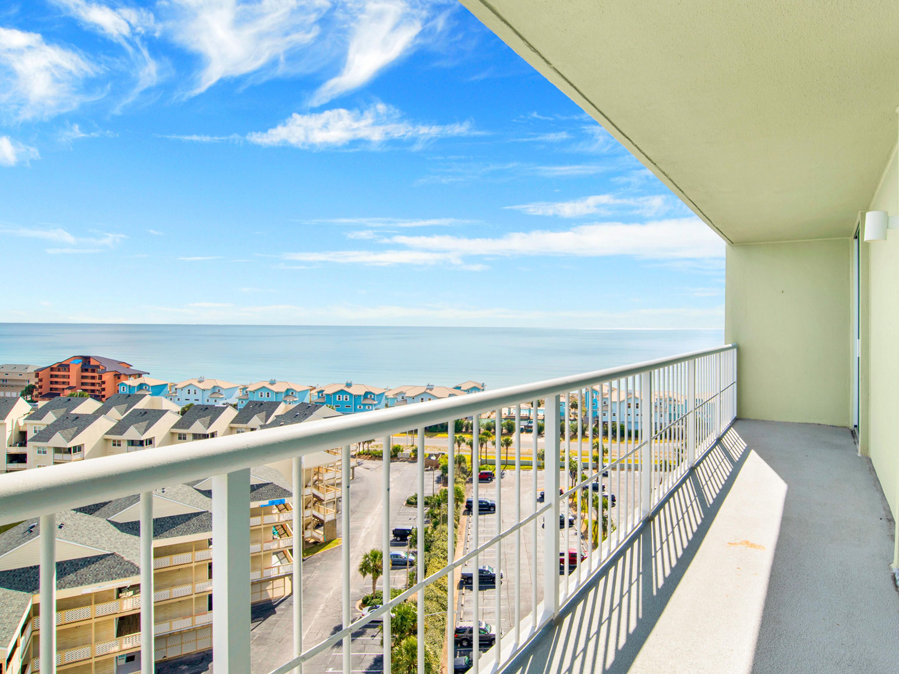 A balcony with a view of a coastal town, including a parking area and buildings with multiple stories.
