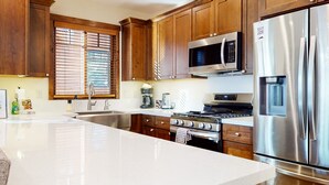 A modern kitchen with wooden cabinets, stainless steel appliances, a large island, and a window with blinds. - Pet-Friendly 5-Star Townhome, Private Hot Tub, Gas Grill, Walk to The Village (Mammoth Lakes)