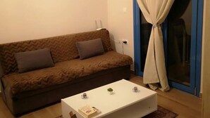A living room with a brown sofa, two pillows, a white coffee table with a book and a small plant, and a window with blue curtains. - 1 Bed Apt - Sleeps 2 - Free Parking - Sea View (Kalymnos)