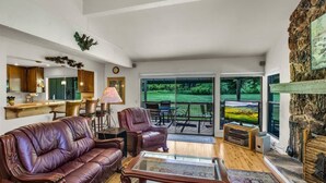 A spacious living room with a leather sofa, a glass coffee table, and a fireplace. There is a dining area with a table and chairs, and a view of a green landscape through large windows. - Views of the 8th Fairway at Plumas Pines-8 Aspen (Blairsden)