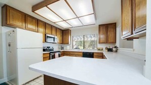 Una cocina con armarios de madera, encimera blanca, microondas integrado y ventana con persianas.