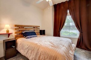 Un dormitorio con cabecera de madera, cama con colcha color beige, mesita de noche con lámpara y ventana con cortinas marrones.