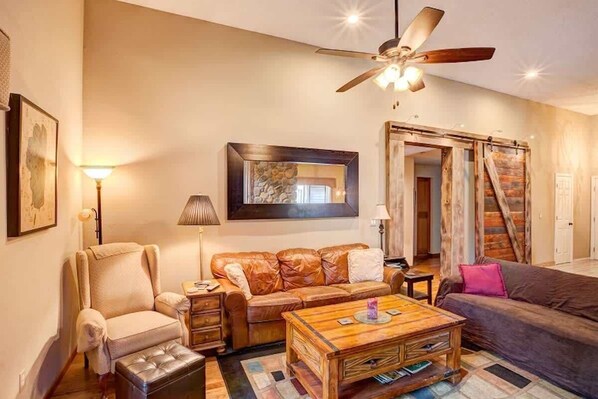 Una sala de estar con ventilador de techo, un sofá de cuero, una mesa de centro de madera y un sillón con reposabrazos acolchados.